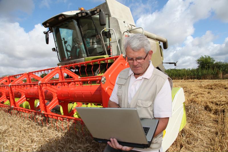 Farmer checking his laptop with farming equipment behind him.